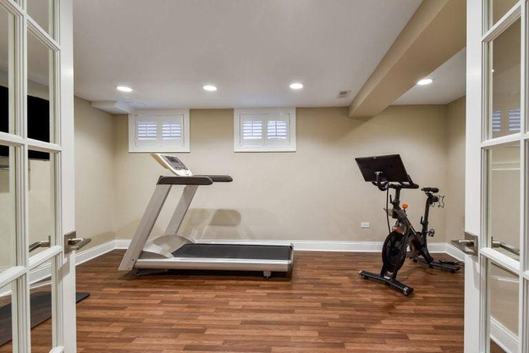 A home gym with an EZ Breathe (Taller Height), Basement Model A-400 – Standard chute – for walls up to 10′ on the left and a stationary bike on the right, set up on a wooden floor with recessed ceiling lights overhead and windows with blinds on the far wall.