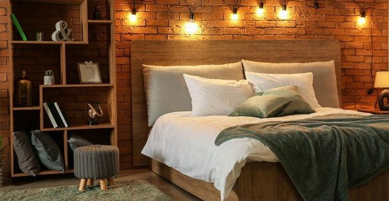 A cozy bedroom with a brick wall, wooden bed frame, white and green bedding, a small ottoman, an EZ Breathe (Taller Height), Basement Model A-400 – Standard chute – for walls up to 10′ shelving unit. String lights are hung above the bed.